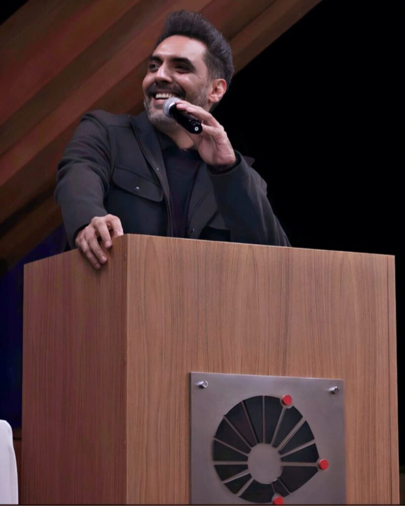 Um homem de pele média, com cabelos escuros e um sorriso amigável está em um palco, atrás de uma tribuna. Ele usa uma jaqueta escura e está falando ao microfone. A tribuna é de madeira clara, com um painel metálico em formato circular no centro da mesa, contendo um padrão geométrico escuro, formando a logomarca da Unicamp. A iluminação e o fundo sugerem um ambiente interno formal de um auditório.