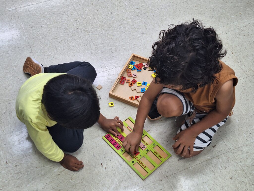Fotografia de dois meninos sentados no chão da Biblioteca da Faculdade de Educação da UNICAMP brincando com um jogo de operações numéricas. O garoto da esquerda é pardo, tem cabelo preto e liso, veste uma blusa de manga comprida na cor verde clara, veste uma calça na cor cinza escuro e usa tênis. O garoto da direita é pardo, tem o cabelo castanho escuro e encaracolado, veste uma blusa regata na cor caramelo, veste uma bermuda branca com listras pretas e usa tênis. Fim da descrição.