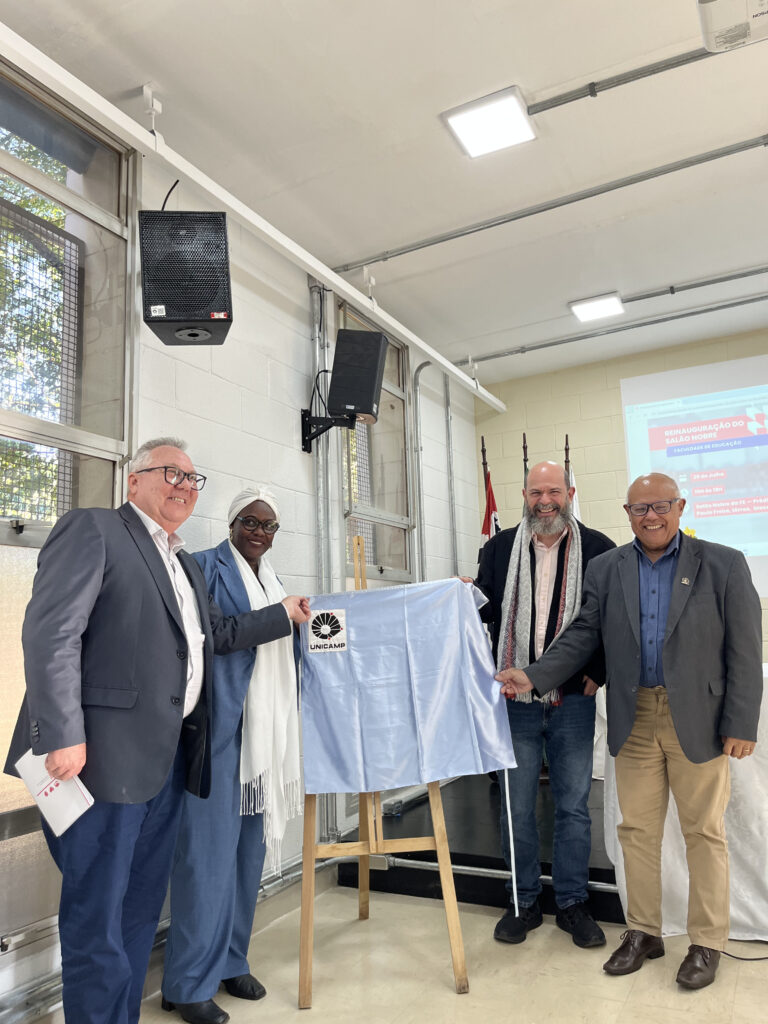Photograph showing four people in a well-lit room. In the center is an easel with a light blue satin covering its contents. The four people point to the easel. There are two white men, a black woman, and a yellow man. All are wearing formal clothes and smiling. End of description.