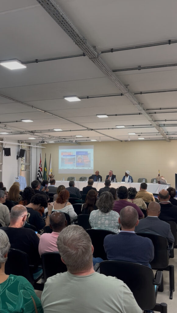 Photograph of a room full of people. In front, there's a themed table with people and a large screen broadcasting images. The space is enclosed, with light-colored walls and ceiling, and is well-lit. End of description.