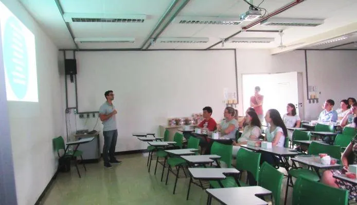 Uma sala de aula com cadeiras universitárias verdes dispostas em fileiras, onde uma pessoa apresenta conteúdo em projetor enquanto outras pessoas assistem sentadas, com duas pessoas em pé ao fundo próximas à porta.
